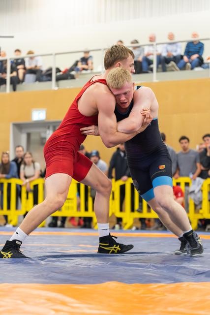 Two young wrestlers locked in a fierce clinch on the mat, both straining with effort, spectators watching from behind yellow barriers.