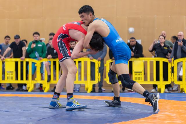 Two wrestlers locked in a fierce grip on the mat, blue singlet athlete dominating red opponent before a crowd.
