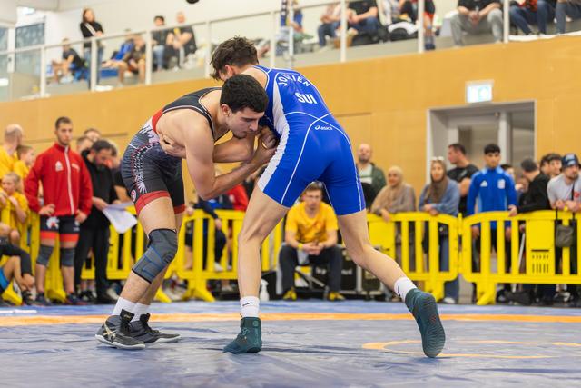 Two wrestlers locked in a fierce grapple on the mat, blue SUI singlet athlete driving forward with determination.