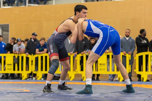 Two wrestlers grip each other intensely on a blue mat, surrounded by spectators behind yellow barriers.