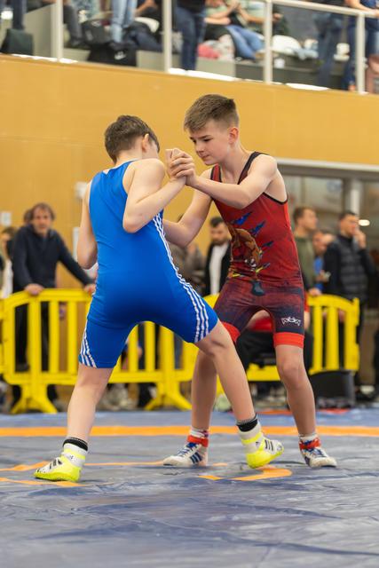 Two young wrestlers grip each other's hands in a tense standoff on the blue mat, spectators watching behind.
