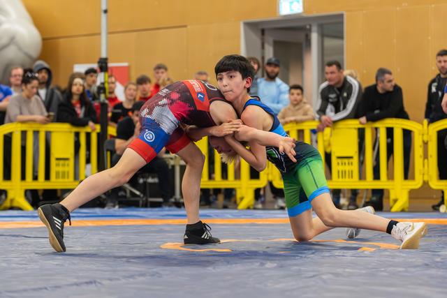 Two young wrestlers locked in a tight grip on the mat, both straining forward with fierce determination during a youth bout.
