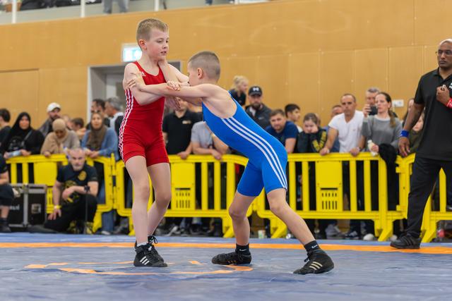 Two young boys lock wrists in an intense standup grapple on a blue mat, watched by a crowded audience.