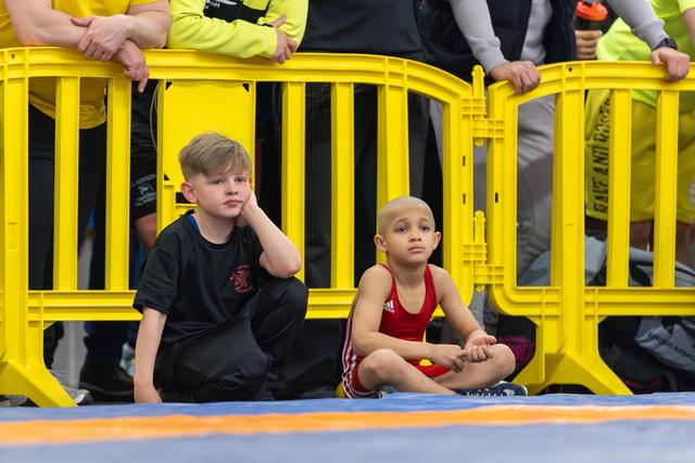 Two young boys sit cross-legged on the mat beside yellow barriers, watching the event with focused, serious expressions.