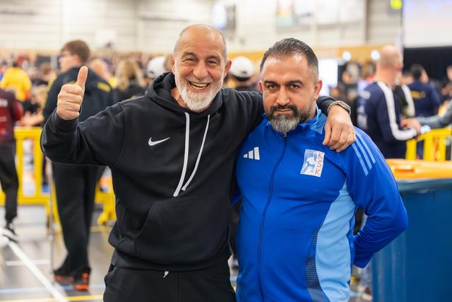 Two coaches smile broadly together; the older man in a Nike hoodie gives a thumbs-up, arm around his colleague in a blue KSK jacket.