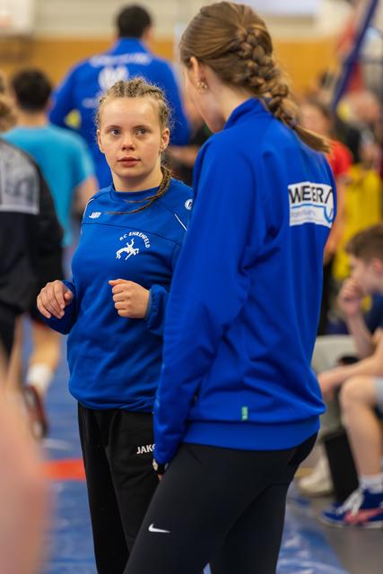 Two young female athletes in blue JAKO jackets talk intently on the gym floor, one facing the camera with focused expression.