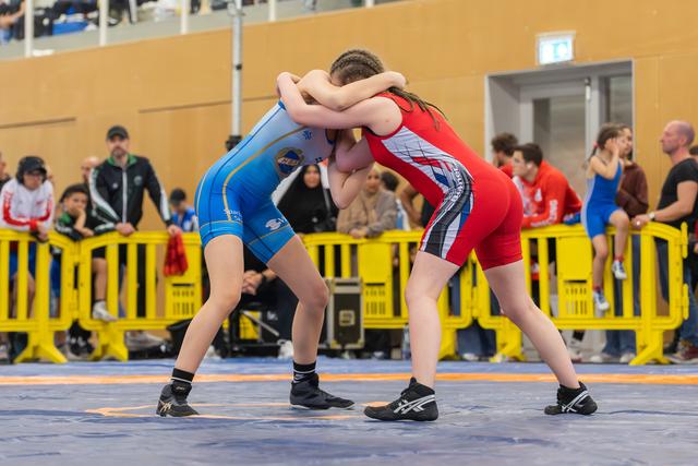 Two young female wrestlers locked in a tight clinch, arms wrapped around each other's necks, intense focus on the mat.