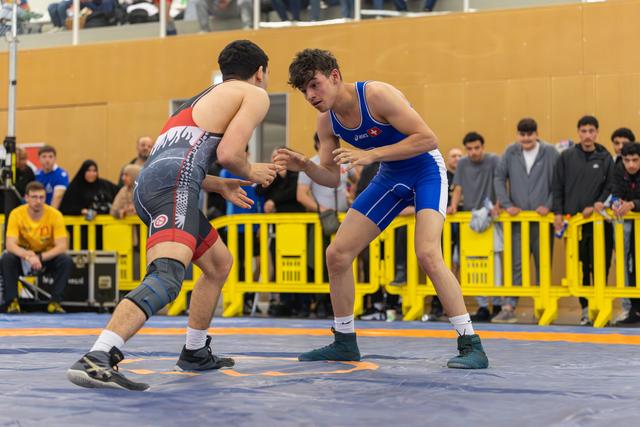 Two young wrestlers face off in a tense stance on the mat, crowds watching behind yellow barriers.