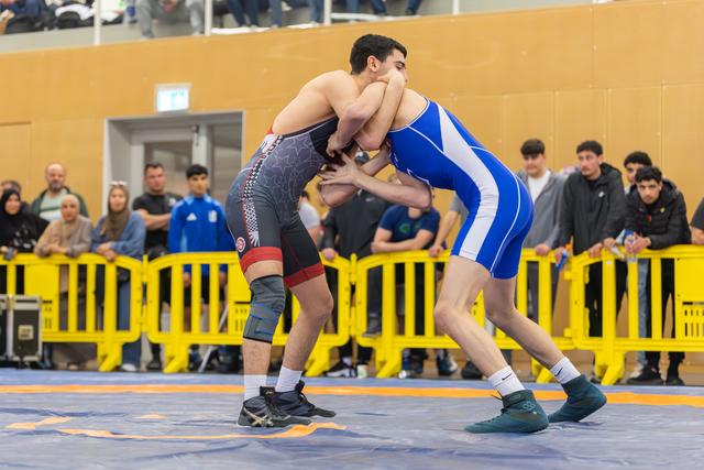 Two male wrestlers lock in a tight clinch on a blue mat, spectators watching intently behind yellow barriers.