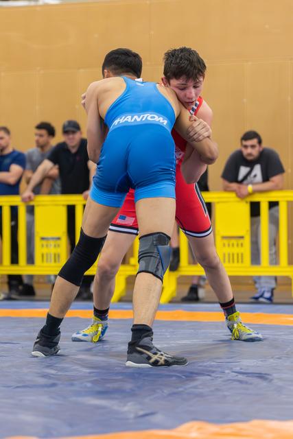 Two young wrestlers locked in a tight clinch on a blue mat, the red-singlet athlete straining with focused intensity.