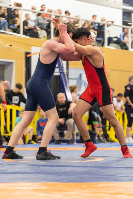 Two wrestlers in navy and red singlets lock arms in an intense clinch on a blue competition mat, crowd watching.