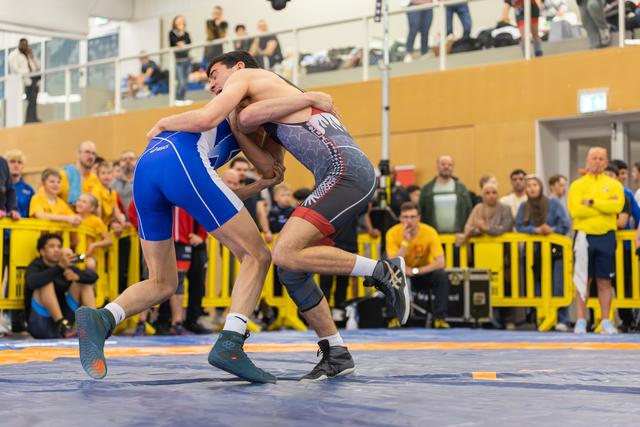 Two wrestlers locked in a tight clinch on the mat, straining against each other with focused intensity before a packed crowd.