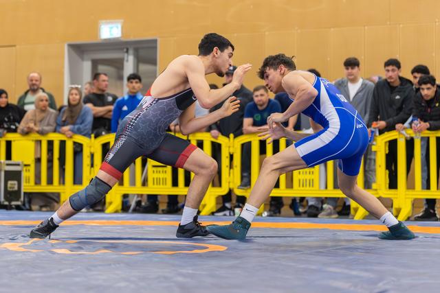 Two young wrestlers in a tense standoff on the mat, one in red-black, one in blue, crowd watching behind yellow barriers.
