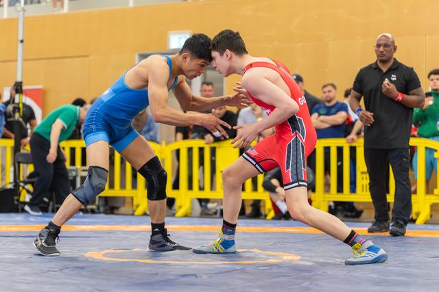 Two young wrestlers in blue and red singlets crouch in intense standoff, hands reaching, referee watching nearby.
