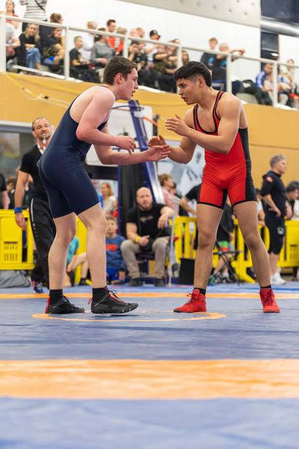 Two teenage wrestlers in navy and red singlets square off, hands extended, intense focus on their faces in a packed arena.