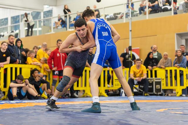 Swiss wrestler in blue singlet locks arms with opponent in red, both low and intense on the mat before a packed crowd.