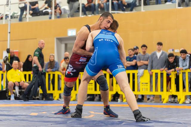 Two wrestlers lock up intensely on the mat; blue SWE BK Velo athlete engages a red-singlet opponent before a crowd.