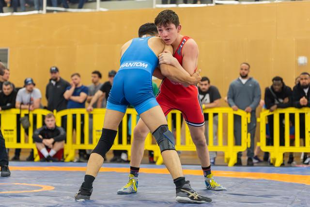 Teen wrestler in red singlet grips opponent in blue, both locked in intense standoff on competition mat.