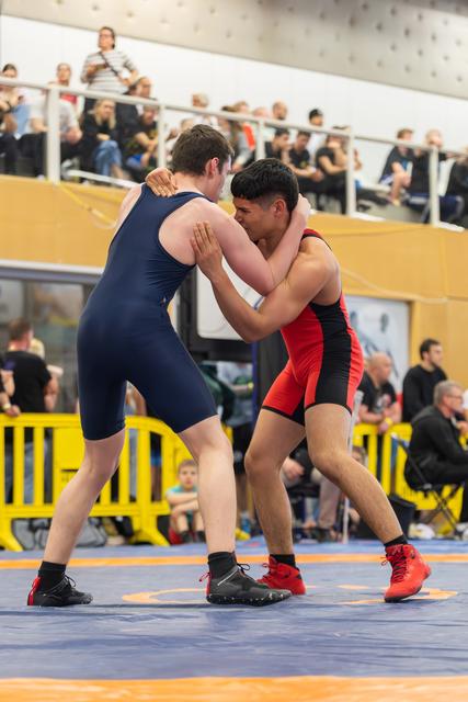 Two young wrestlers in navy and red singlets lock up in a tie position on the mat, focused and competitive.