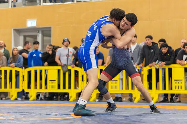 Two wrestlers locked in a tight clinch on the mat, the athlete in blue straining forward with intense focus before a watching crowd.