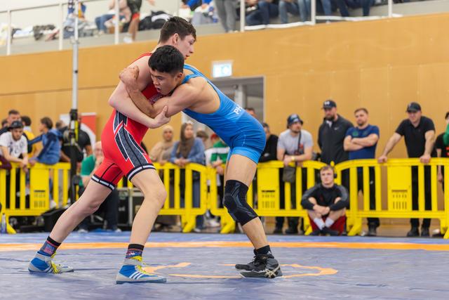 Two young wrestlers locked in a tight clinch on the mat, both leaning forward with intense focus during a competitive bout.