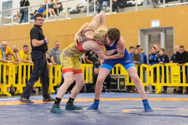 Two young wrestlers locked in a fierce clinch on a blue mat, referee watching closely as crowd looks on.