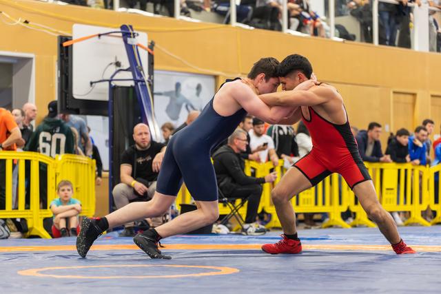Two wrestlers clash forehead-to-forehead in intense clinch, one in navy, one in red, on a blue mat before a crowd.