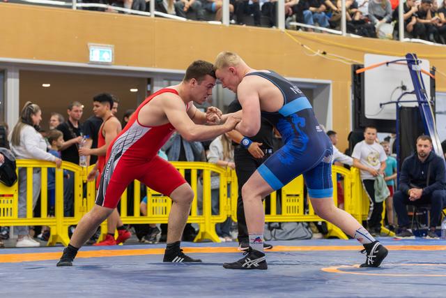 Two wrestlers in red and blue singlets lock hands in an intense face-off on an orange mat, surrounded by spectators.