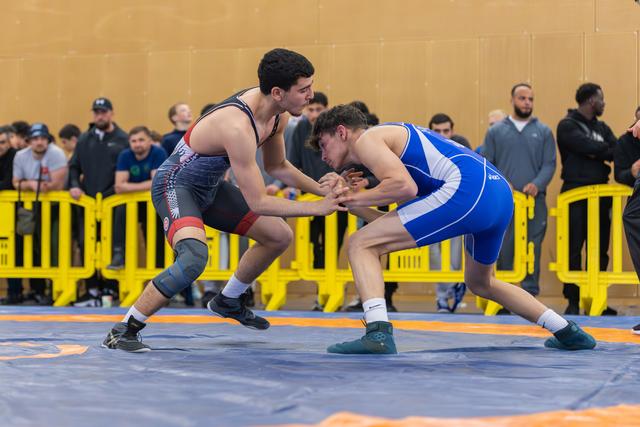Two young wrestlers in black and blue singlets face off in a crouch, hands clasped, intense focus on the blue mat.