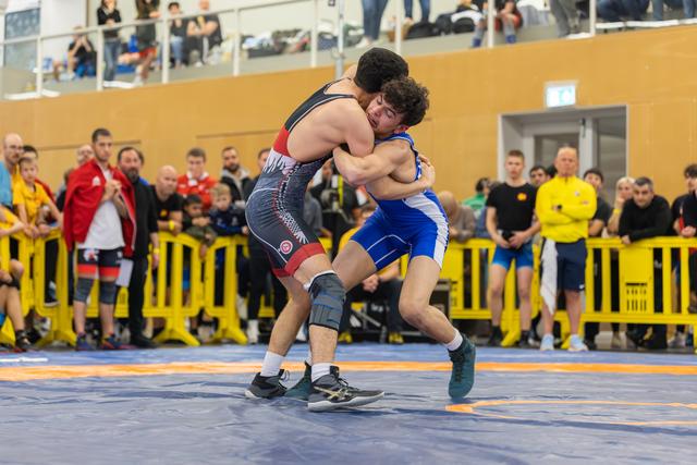 Two wrestlers locked in a fierce clinch on the mat, both straining with effort as spectators watch closely behind barriers.