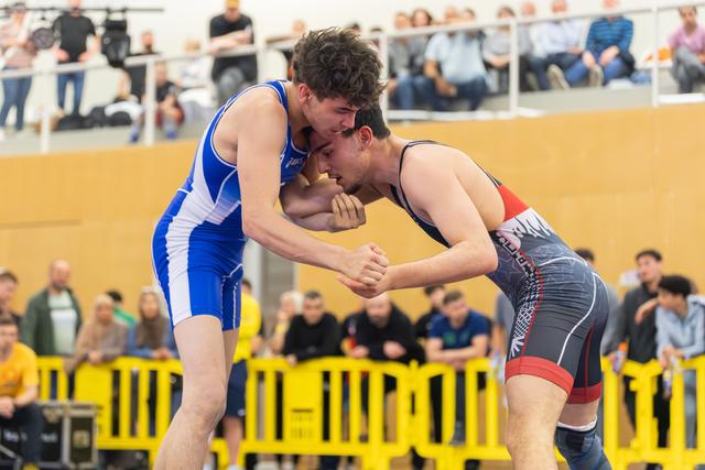 Two young wrestlers lock heads in an intense clinch, both straining with determination on an indoor mat before a crowd.
