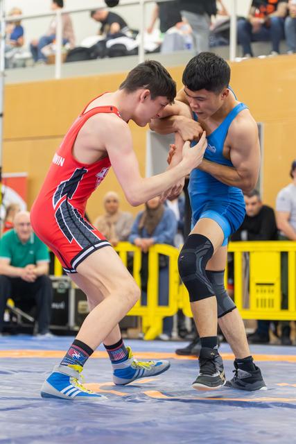 Two young wrestlers locked in a fierce clinch on the mat, red vs blue singlet, spectators watching in background.