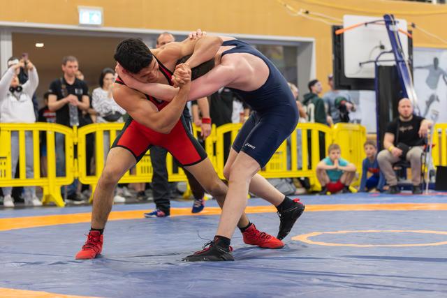 Two wrestlers locked in a fierce clinch on the blue mat, one in red, one in navy, straining for control at a sports hall event.