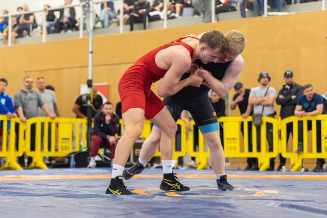 Two wrestlers locked in a powerful clinch on the mat, both leaning forward with focused determination during a competitive bout.