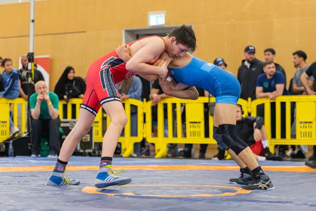 Two young wrestlers locked in an intense clinch on the mat, both straining forward in red and blue singlets.