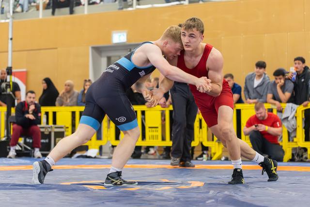 Two male wrestlers in red and blue singlets locked in an intense clinch, both leaning forward with fierce focus on the mat.