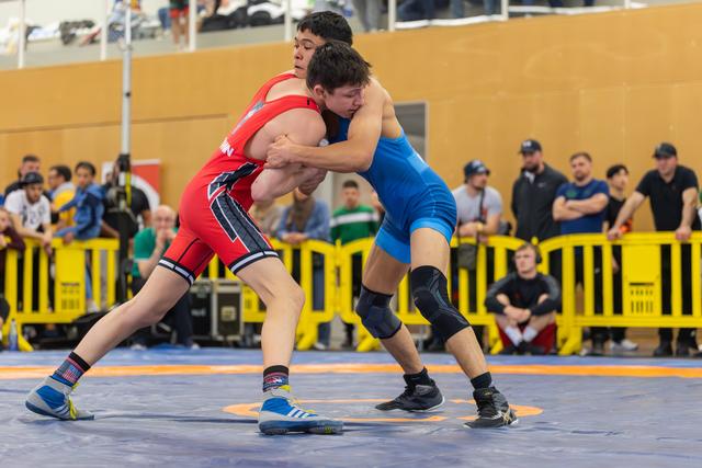 Two young wrestlers lock up in fierce competition, one in red, one in blue, straining forward on a blue mat.