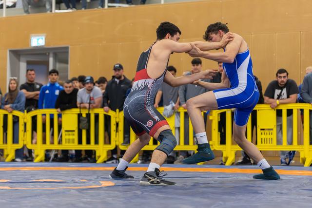 Two young wrestlers locked in a standing clinch on a blue mat, both pushing aggressively, crowd watching behind yellow barriers.