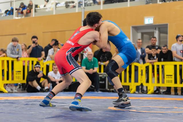 Two wrestlers locked in a fierce clinch on the mat, one in red, one in blue, crowds watching behind yellow barriers.