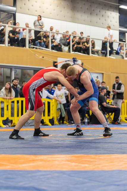 Two male wrestlers locked in a tense grapple, both leaning forward aggressively on a blue and orange mat before a crowd.