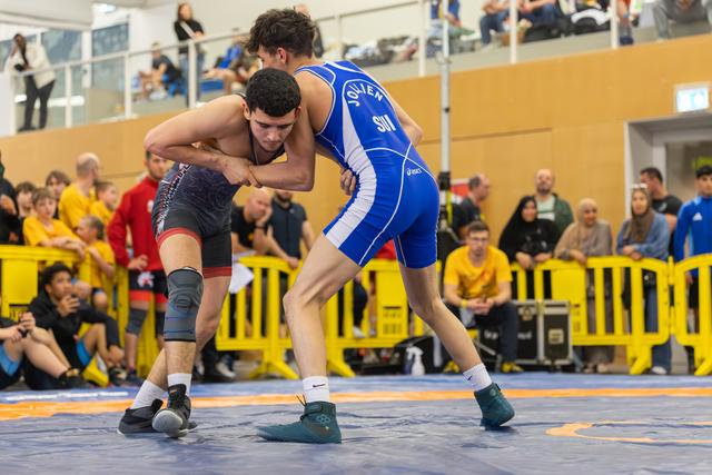 Two young wrestlers lock grips in tense standoff on blue mat, crowd watching closely in packed indoor venue.