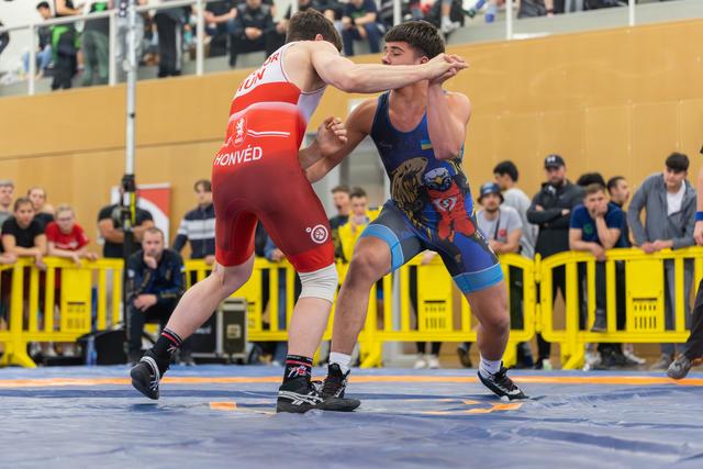 Two wrestlers in red and blue singlets battle on the mat, both straining with focused intensity as spectators watch behind barriers.