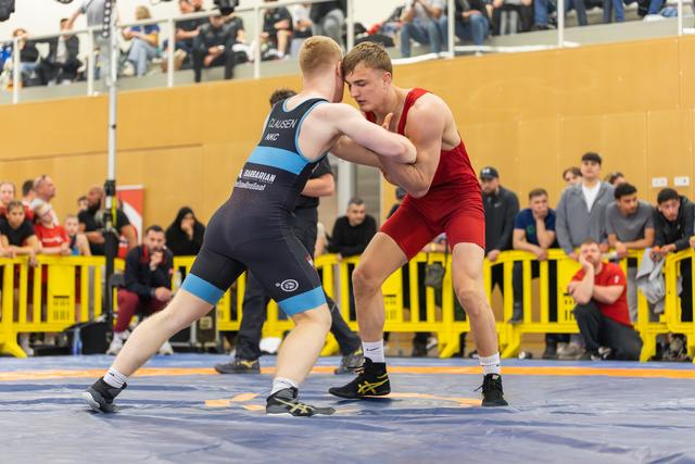 Two young wrestlers in blue and red singlets face off in a tense, crouched stance on a blue competition mat, crowd watching.