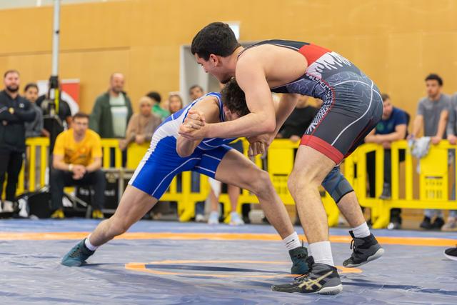 Two male wrestlers in intense clinch, blue singlet athlete defending against black-red opponent on blue mat, crowd watching.