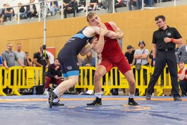 Two young wrestlers lock arms on a blue mat, the red-clad athlete pushing forward with intense focus during a competitive bout.