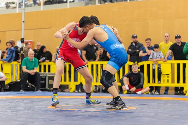 Two young wrestlers lock grips intensely on the mat, one in red, one in blue, surrounded by spectators.