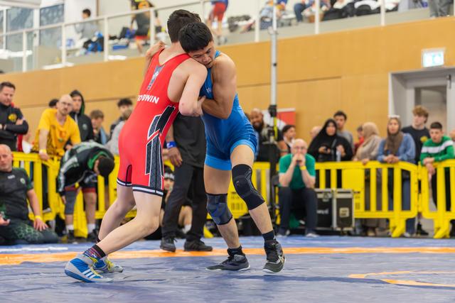 Two young wrestlers locked in a tight clinch on the mat, straining for control in front of a packed crowd.