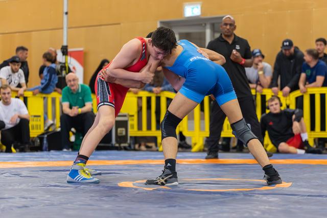 Two young wrestlers lock up in an intense clinch on the mat, red vs blue, surrounded by spectators in an indoor venue.
