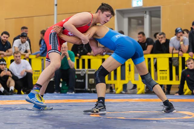 Two young wrestlers locked in a fierce grip on the mat, red vs blue singlets, crowds watching behind yellow barriers.