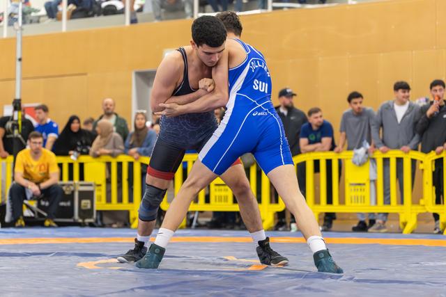 Swiss wrestler in blue singlet grips opponent in dark uniform, both locked in tense struggle on blue mat before crowd.